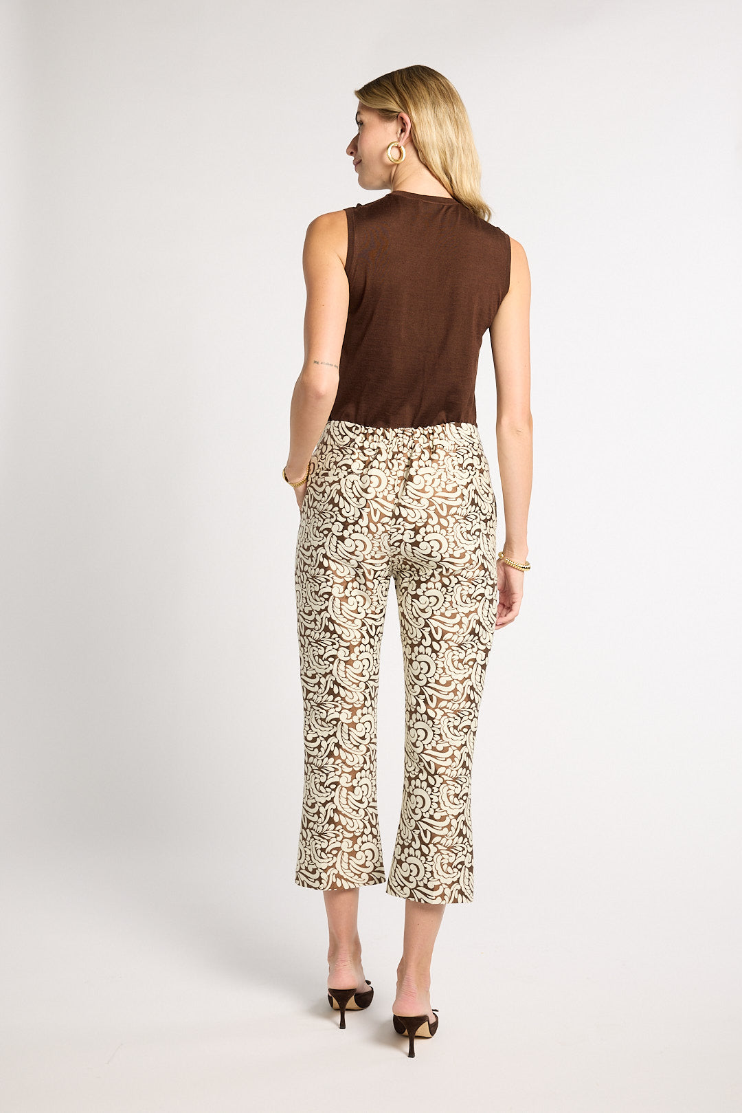 A woman shown from the back wearing a sleeveless brown top and patterned cropped pants with an abstract floral print in beige, brown, and black, paired with black heels, against a light gray studio background.