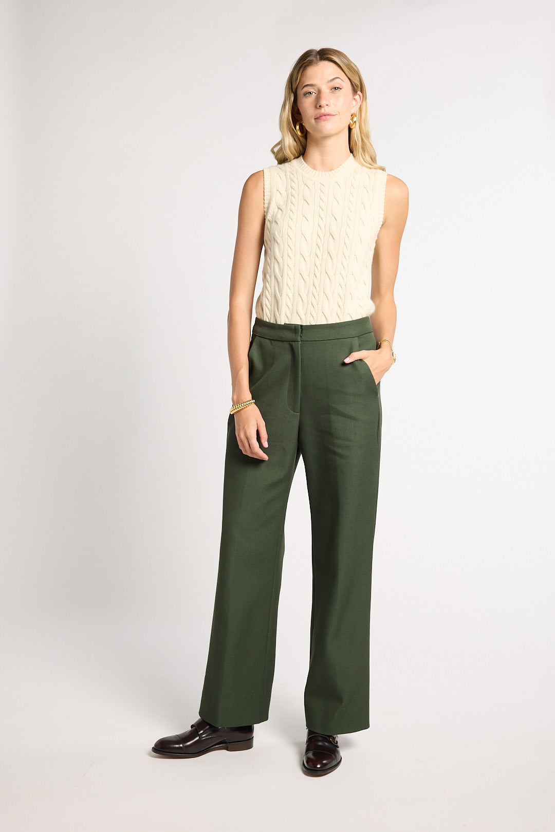 A woman modeling a sleeveless cream cable-knit top and dark olive green wide-leg trousers, paired with dark brown leather shoes, against a light gray studio background.