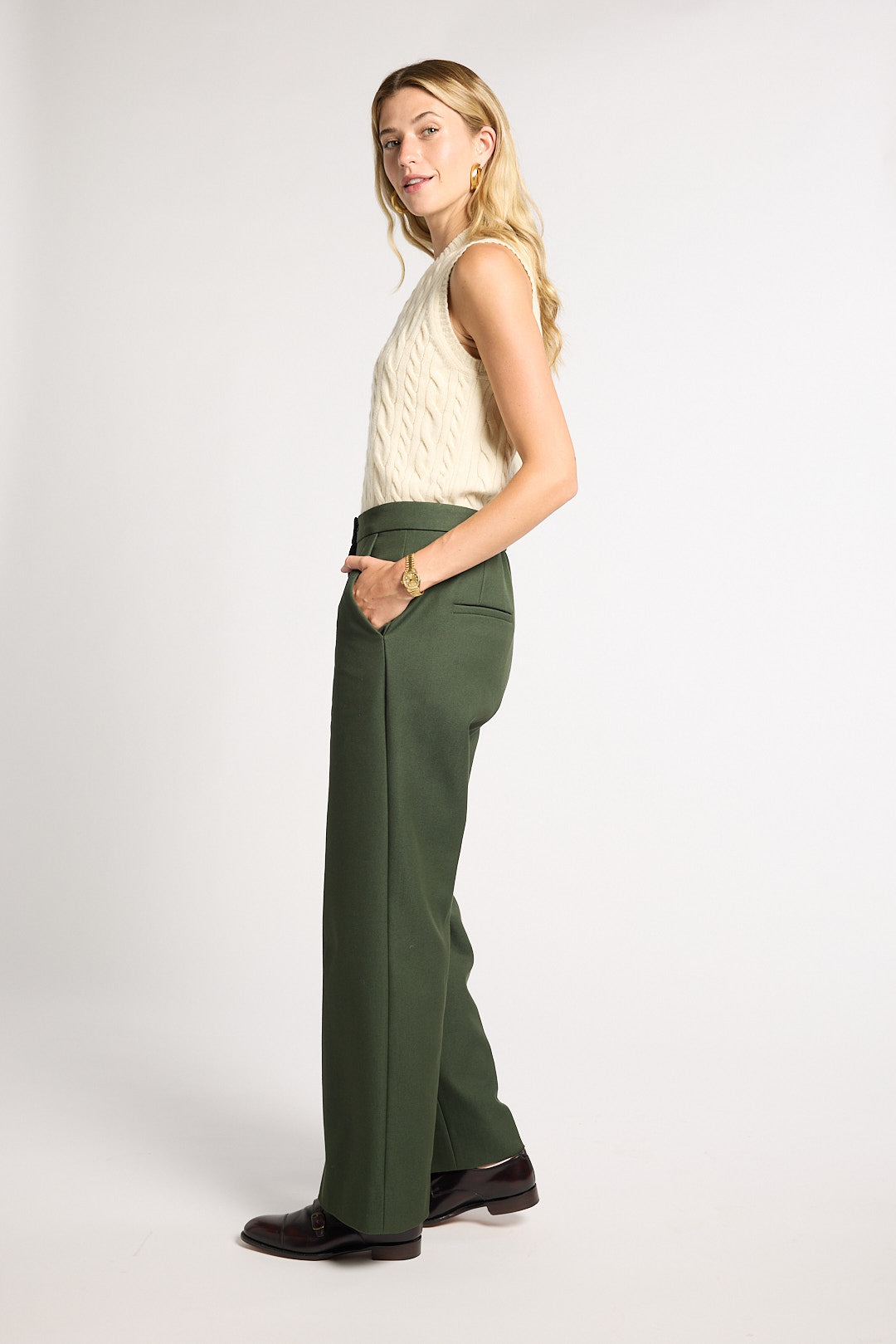 A woman modeling a pair of high-waisted, wide-leg olive green trousers, paired with a cream cable-knit sleeveless top and dark brown leather loafers, against a plain light gray studio background.