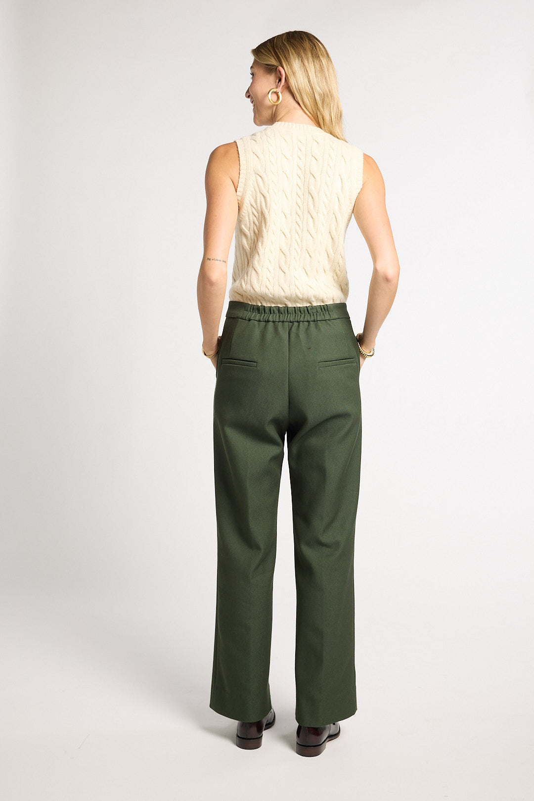 A woman is shown from the back wearing a cream cable-knit sleeveless top and dark olive green wide-leg trousers, standing against a light gray studio background.