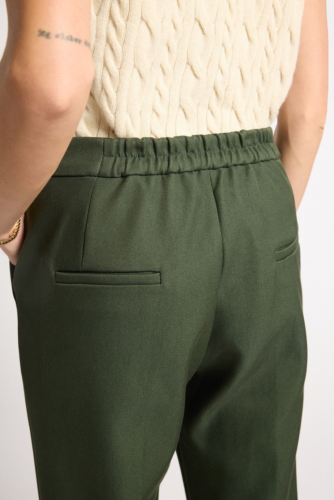 A close-up back view of a person wearing olive green trousers with an elastic waistband and side pockets, paired with a cream cable-knit top against a plain light background.