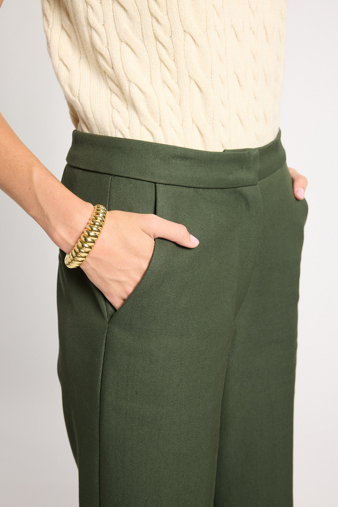 A close-up of a person wearing dark olive green wide-leg trousers with a cream cable-knit top. The person's hand is in a pocket, wearing a chunky gold chain bracelet, against a light gray background.