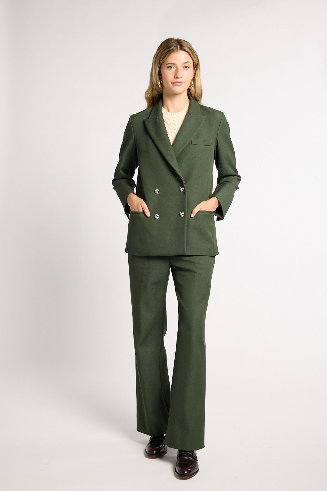A woman modeling a dark green double-breasted blazer and matching wide-leg trousers, styled with a cream top and dark brown loafers, against a light gray studio background.
