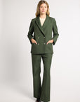 A woman modeling a dark green double-breasted blazer and matching wide-leg trousers, styled with a cream top and dark brown loafers, against a light gray studio background.