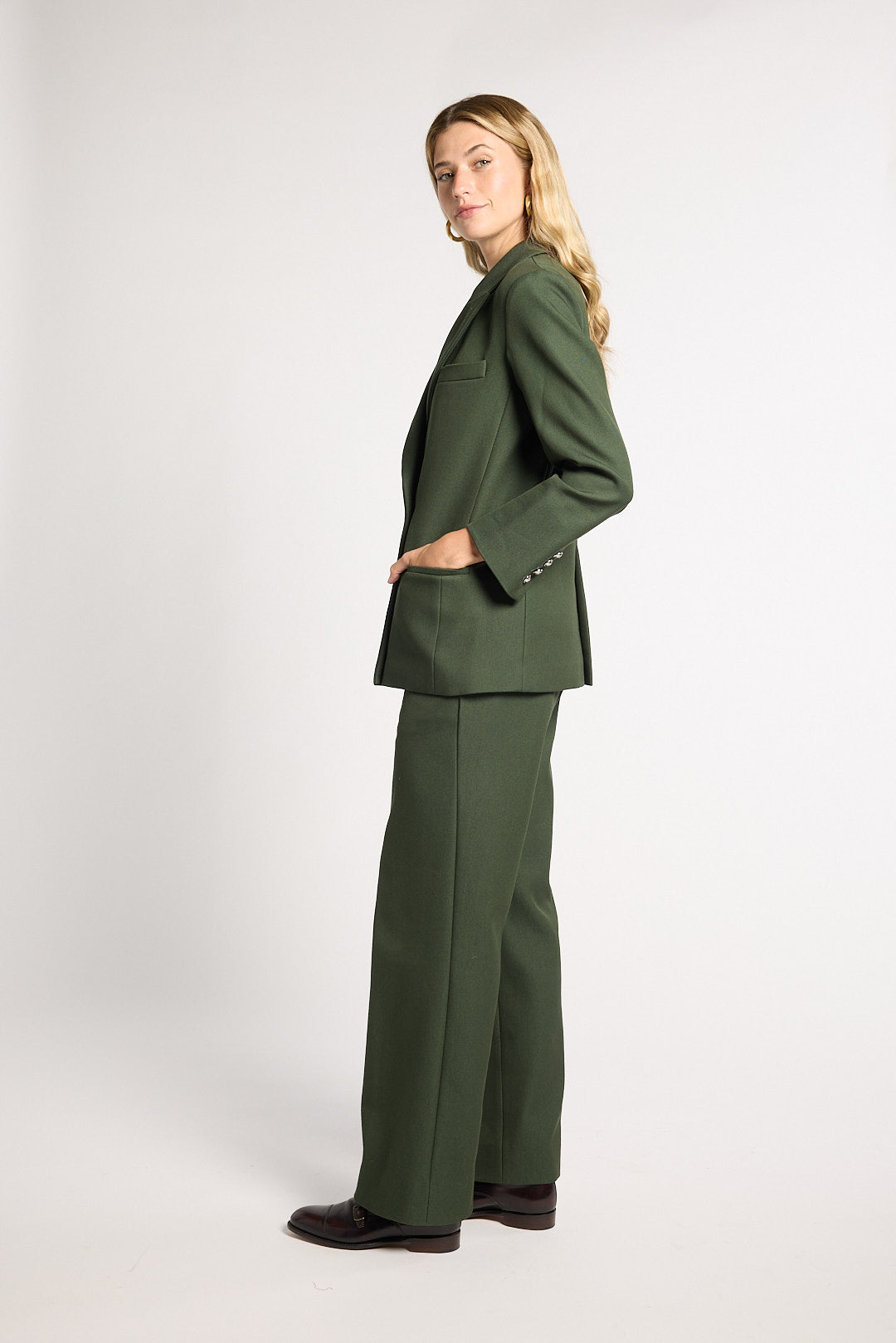 A woman modeling a dark green tailored pantsuit, featuring a single-breasted blazer with structured shoulders and matching wide-leg trousers, photographed against a neutral light gray studio background.
