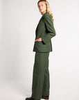 A woman modeling a dark green tailored pantsuit, featuring a single-breasted blazer with structured shoulders and matching wide-leg trousers, photographed against a neutral light gray studio background.