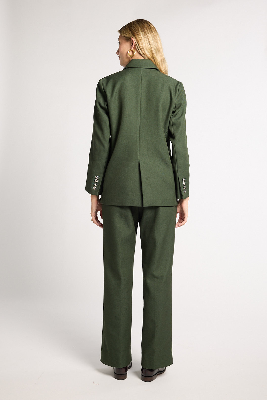 A woman shown from the back wearing a matching olive green suit, featuring a structured blazer with button details on the cuffs and wide-leg trousers, set against a plain light gray studio background.
