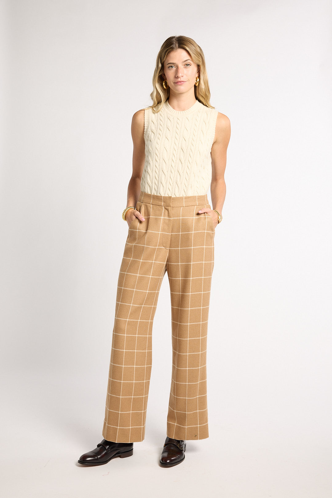 A woman modeling a sleeveless cream cable-knit top and tan windowpane plaid wide-leg trousers with black leather loafers, shot against a light gray studio background.