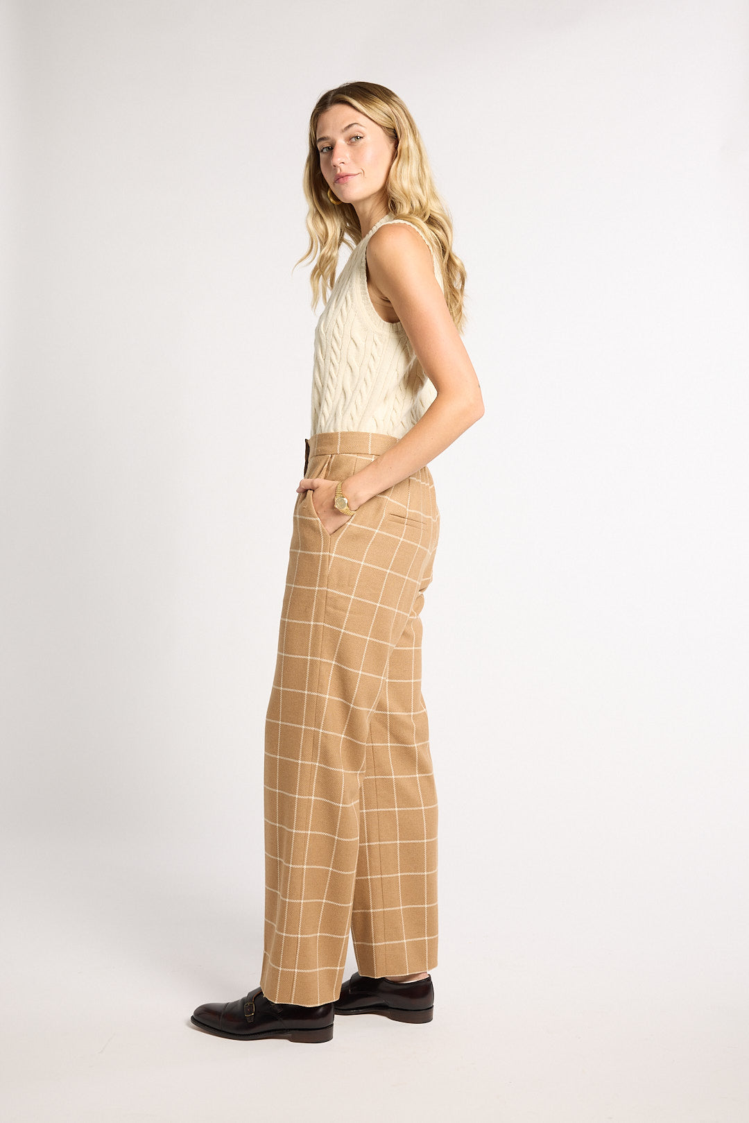 A woman stands in profile wearing a cream sleeveless knit top and tan wide-leg trousers with a white windowpane plaid pattern, paired with black shoes, against a plain light gray studio background.