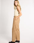 A woman stands in profile wearing a cream sleeveless knit top and tan wide-leg trousers with a white windowpane plaid pattern, paired with black shoes, against a plain light gray studio background.