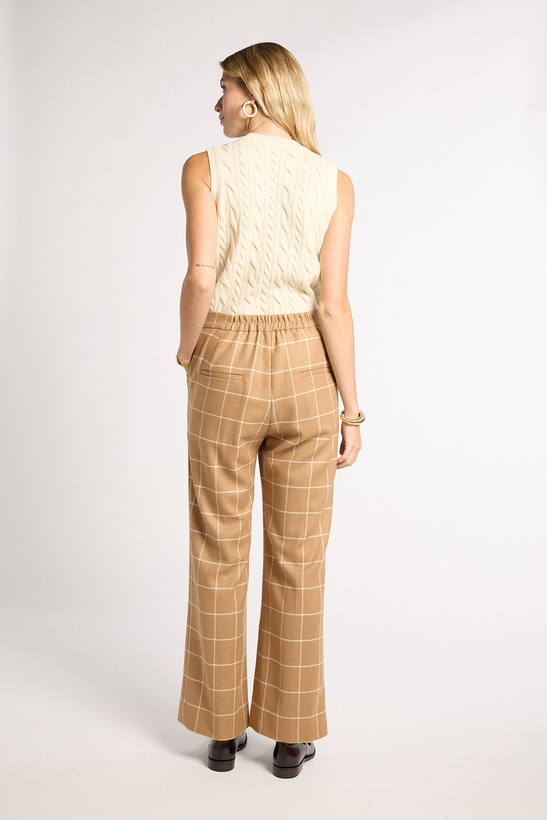 A woman is shown from the back wearing tan wide-leg trousers with a white windowpane check pattern and a cream cable-knit sleeveless top, standing against a light gray studio background.