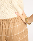 Close-up of a person wearing a cream cable-knit sweater and tan windowpane plaid pants, with one hand in the pocket and gold beaded bracelets on the wrist, against a plain white background.
