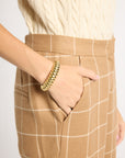 A close-up of a person wearing tan windowpane plaid trousers and a cream cable-knit sweater, with one hand in a pocket and wearing a chunky gold bangle.