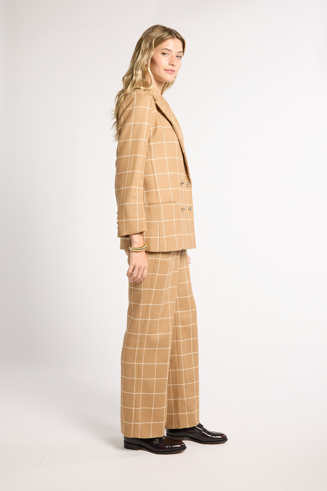 A woman modeling a beige windowpane plaid blazer and matching wide-leg trousers with black leather shoes, standing in profile against a light gray studio background.