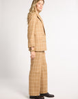 A woman modeling a beige windowpane plaid blazer and matching wide-leg trousers with black leather shoes, standing in profile against a light gray studio background.