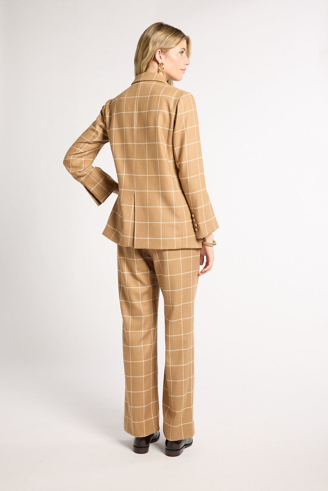 A woman shown from the back wearing a camel-colored two-piece suit with a windowpane plaid pattern, photographed against a plain light gray studio background.