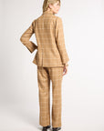 A woman shown from the back wearing a camel-colored two-piece suit with a windowpane plaid pattern, photographed against a plain light gray studio background.