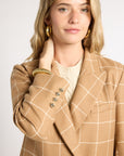 A woman wearing a tan windowpane plaid blazer over a cream top, accessorized with gold hoop earrings and a gold beaded bracelet, photographed against a light gray studio background.