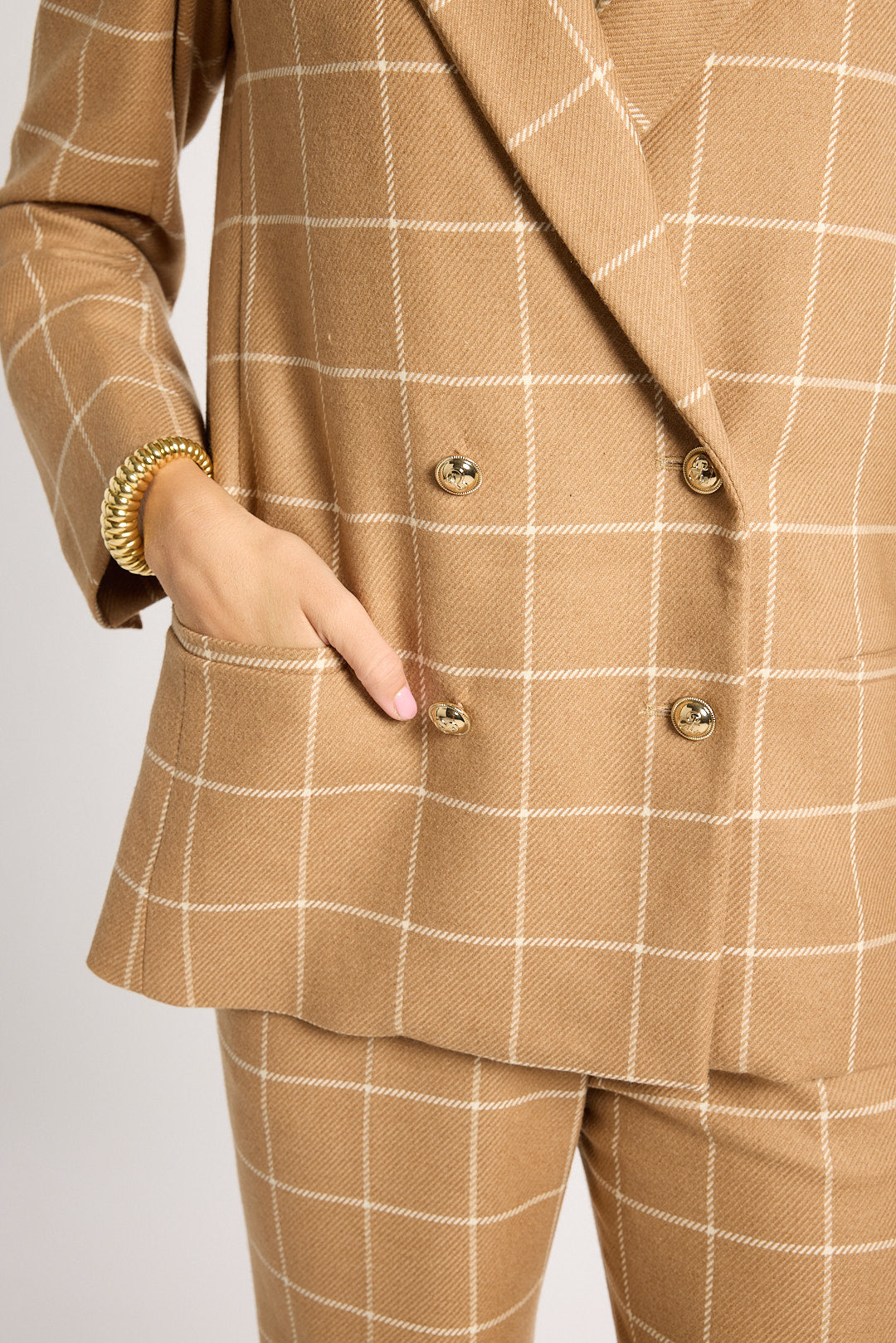 A close-up of a person wearing a tan double-breasted blazer and matching trousers with a white windowpane check pattern. The blazer features gold buttons and a pocket with the person's hand inside.