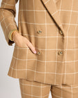 A close-up of a person wearing a tan double-breasted blazer and matching trousers with a white windowpane check pattern. The blazer features gold buttons and a pocket with the person's hand inside.