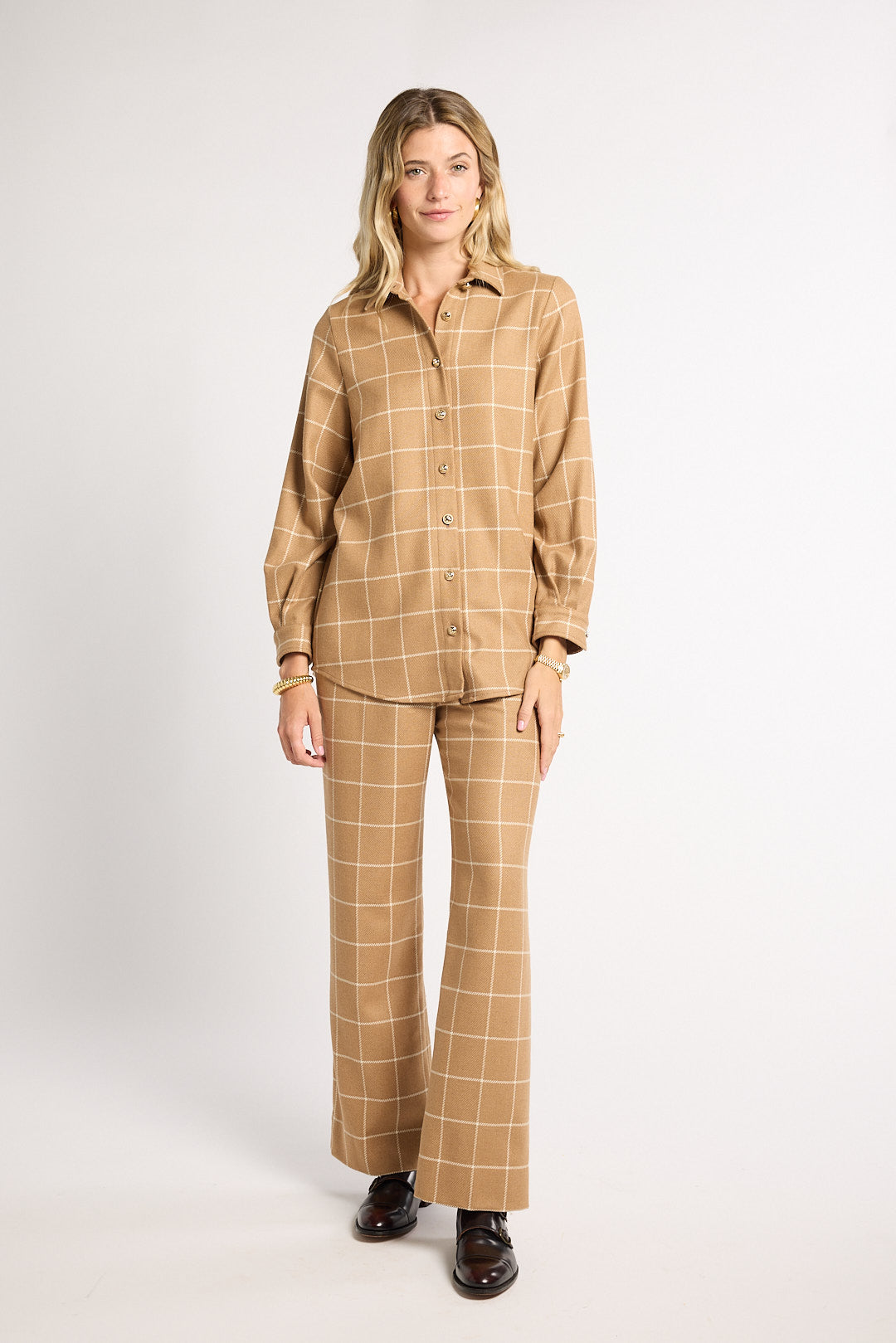 A woman modeling a matching tan and beige plaid two-piece set consisting of a long-sleeve button-up shirt and wide-leg trousers, paired with dark brown leather shoes, against a plain light gray studio background.