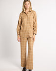 A woman modeling a matching tan and beige plaid two-piece set consisting of a long-sleeve button-up shirt and wide-leg trousers, paired with dark brown leather shoes, against a plain light gray studio background.