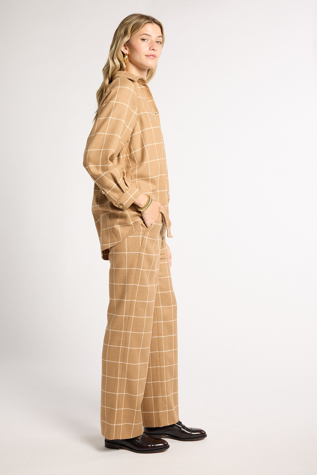 A woman modeling a matching beige and white windowpane plaid lounge set, featuring a long-sleeve top and wide-leg trousers, paired with black leather loafers against a plain white studio background.