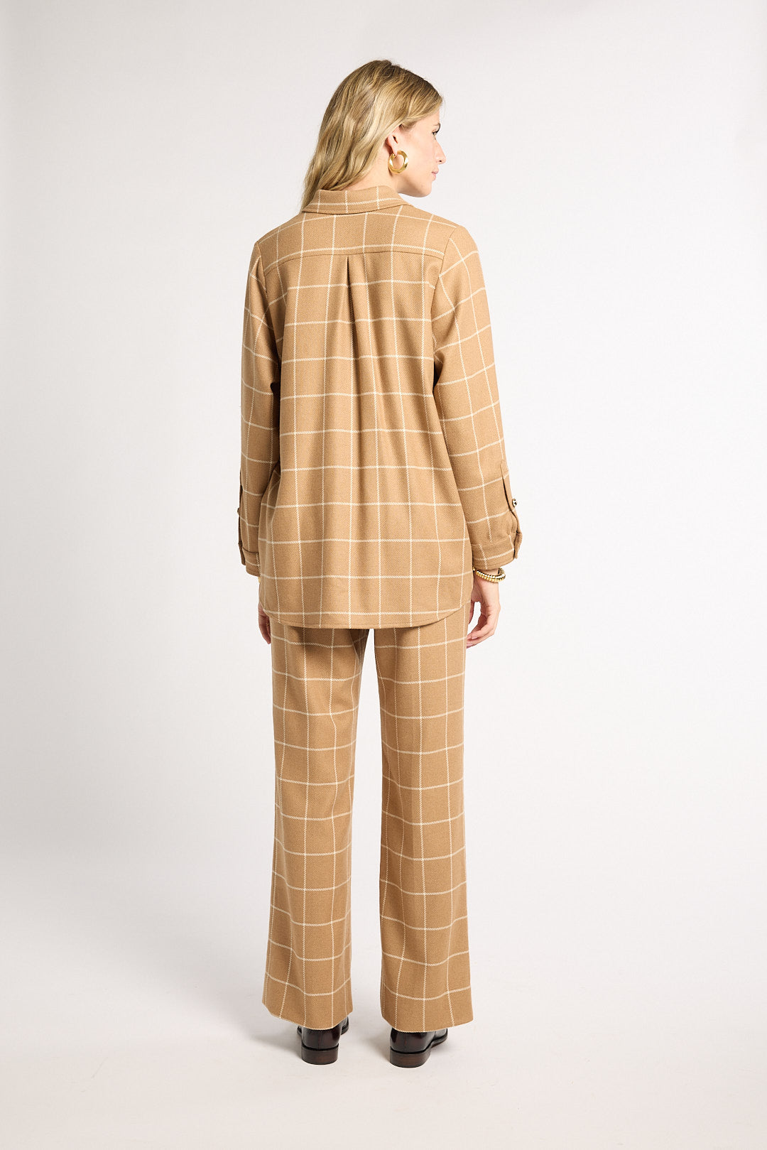 A woman viewed from the back wearing a tan and beige windowpane plaid blazer and matching wide-leg trousers, standing against a light gray studio background.