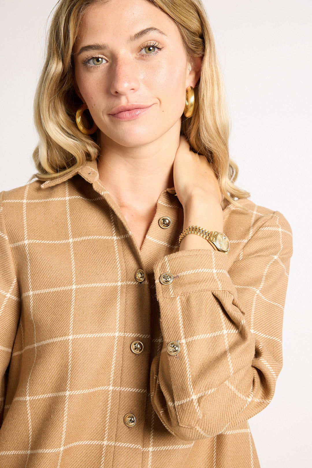 A woman wearing a tan and beige windowpane plaid button-down shirt jacket, styled with gold hoop earrings and a gold watch, against a plain light gray background.