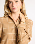 A woman wearing a tan and beige windowpane plaid button-down shirt jacket, styled with gold hoop earrings and a gold watch, against a plain light gray background.