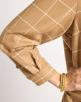 Close-up of a person wearing a tan and white windowpane plaid blazer or overshirt, with a gold beaded bracelet visible on the wrist, against a light gray studio background.