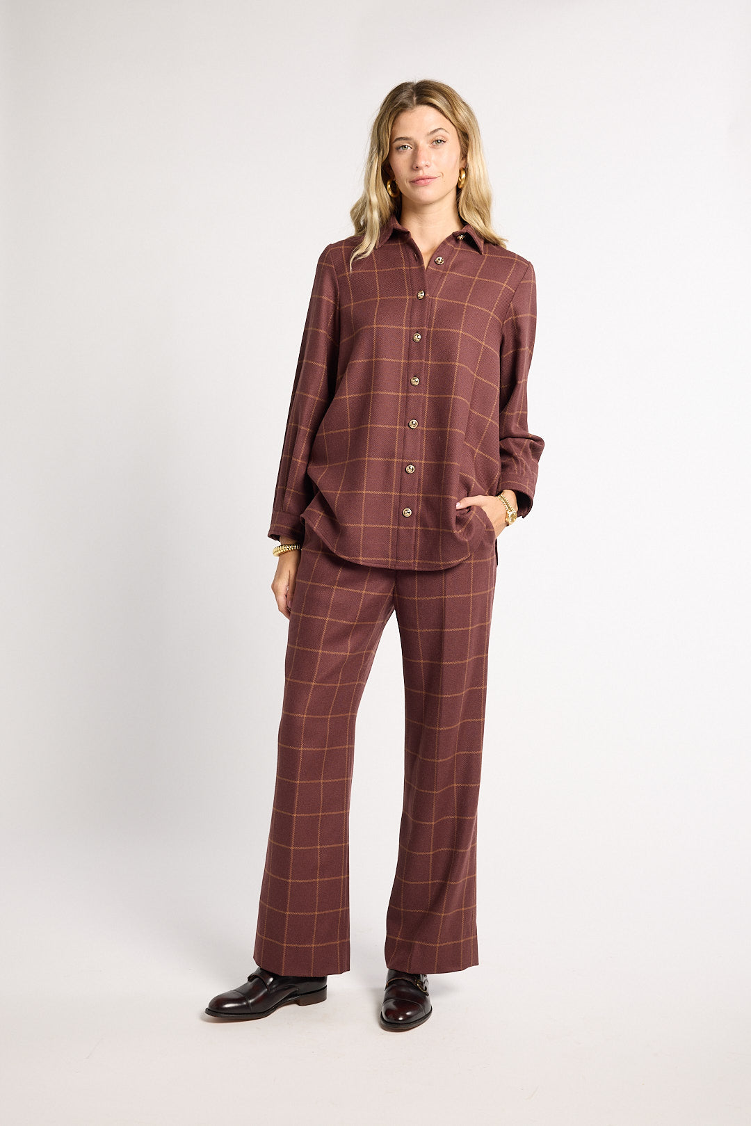 A woman modeling a matching two-piece plaid lounge set in a dusty rose-brown color with thin tan windowpane lines, consisting of a long-sleeve button-up shirt and wide-leg pants, paired with black leather shoes against a light gray studio background.