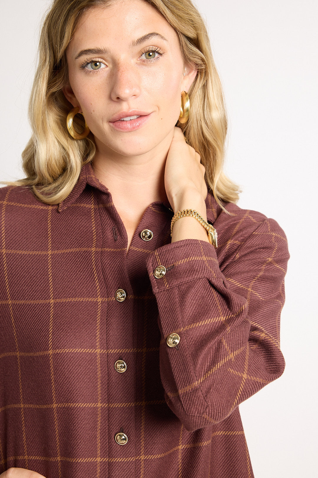A woman wearing a brown and rust plaid button-down shirt with gold buttons, styled with gold hoop earrings and a gold bracelet against a plain white background.