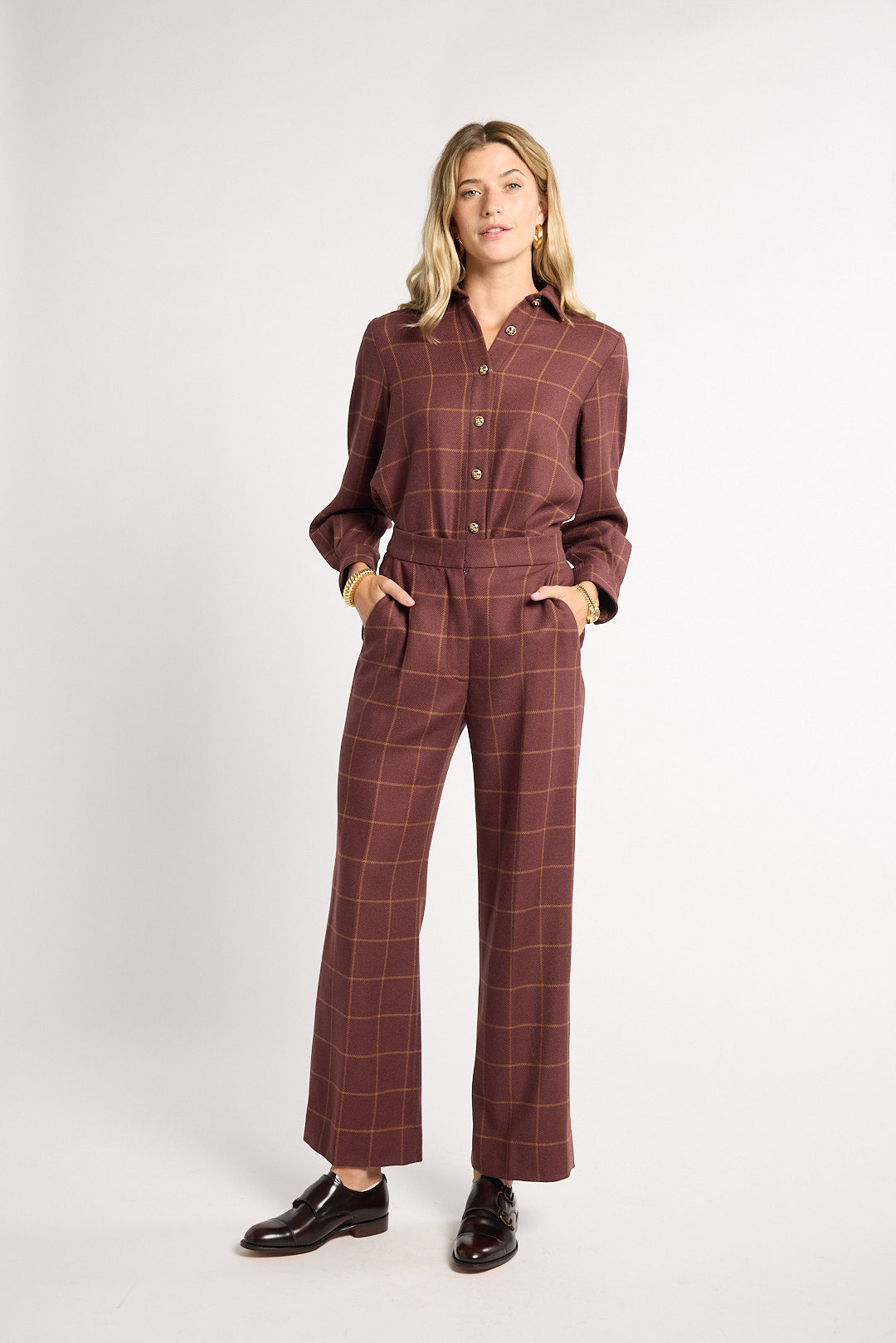 A woman modeling a brown and burgundy windowpane plaid long-sleeve button-up shirt and matching wide-leg trousers, styled with black leather loafers against a plain white studio background.