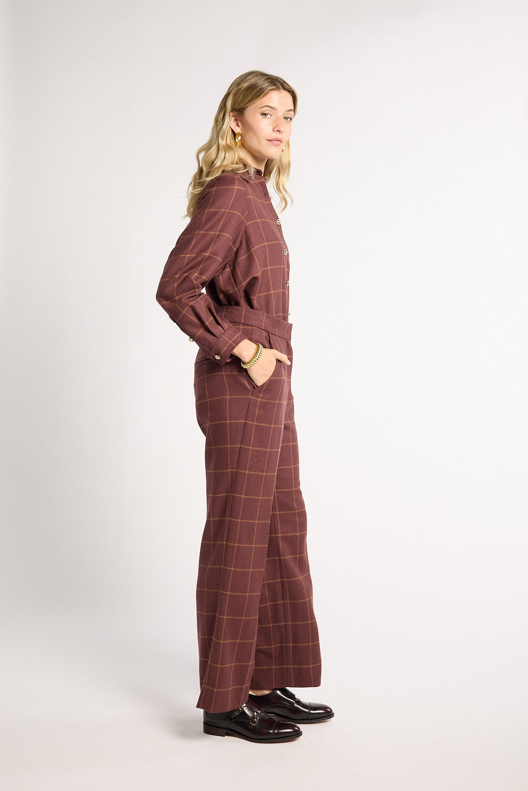 A woman modeling a matching two-piece plaid jumpsuit in a dusty rose-brown color with thin yellow windowpane lines, standing in profile against a light gray studio background.