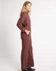 A woman modeling a matching two-piece plaid jumpsuit in a dusty rose-brown color with thin yellow windowpane lines, standing in profile against a light gray studio background.