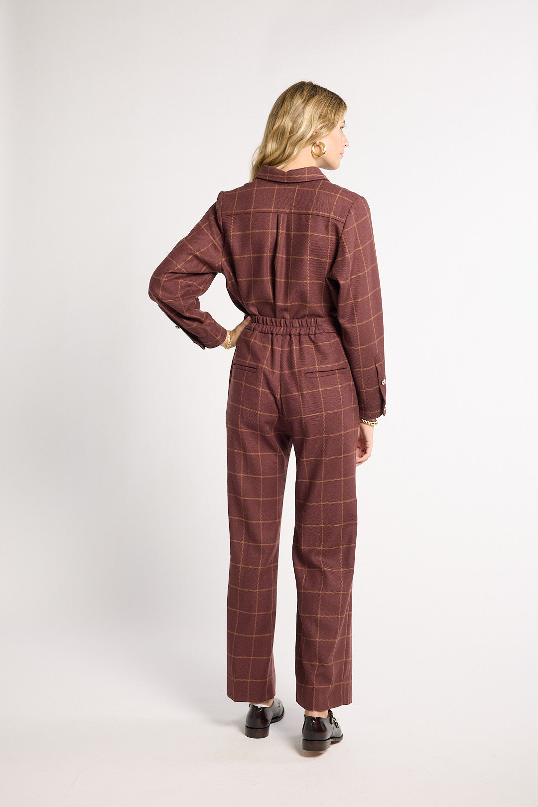 A woman is shown from the back wearing a matching long-sleeve collared jumpsuit in a dark reddish-brown color with a thin windowpane plaid pattern. The jumpsuit has an elastic waist and straight legs, paired with dark brown shoes, against a plain light gray studio background.