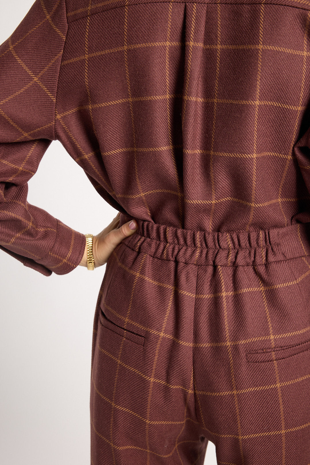 Close-up of a person wearing a brown and gold windowpane plaid jumpsuit, showing the back view with an elastic waistband and pocket details.