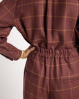 Close-up of a person wearing a brown and gold windowpane plaid jumpsuit, showing the back view with an elastic waistband and pocket details.