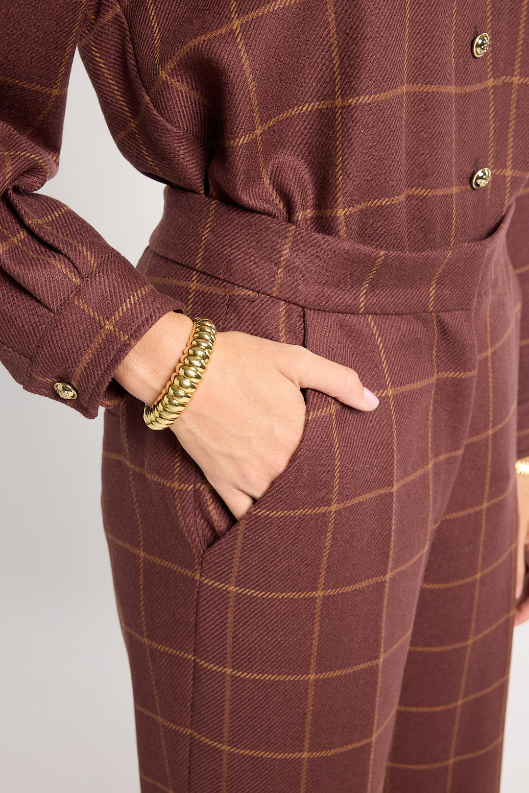 A close-up of a person wearing a brown and tan windowpane plaid jumpsuit, with one hand in a pocket and wearing a chunky gold chain bracelet.