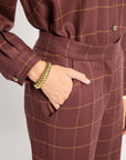 A close-up of a person wearing a brown and tan windowpane plaid jumpsuit, with one hand in a pocket and wearing a chunky gold chain bracelet.