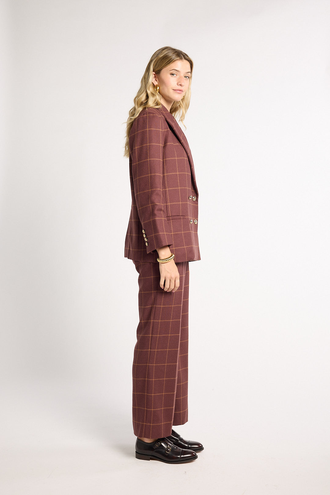 A woman modeling a matching two-piece suit in a dusty rose-brown color with a thin tan windowpane plaid pattern, shown from a side profile against a light gray studio background.