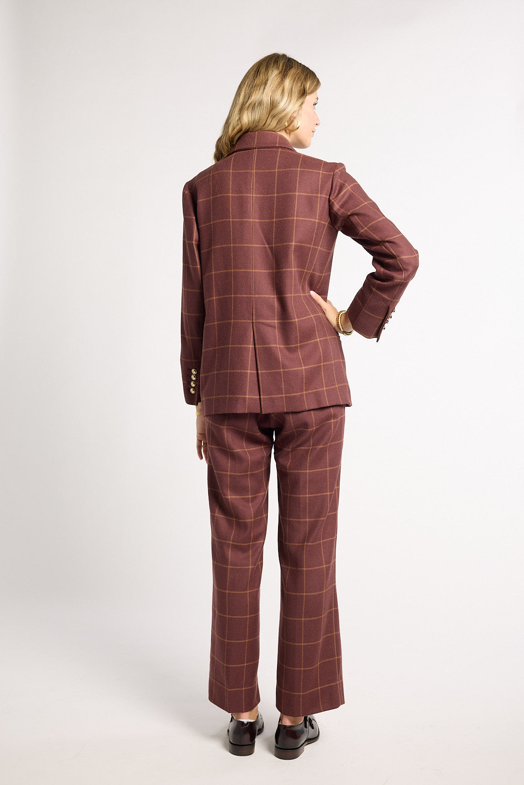 A woman is shown from the back wearing a matching brown and red windowpane plaid blazer and trouser set, paired with black shoes, against a plain light gray studio background.