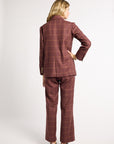 A woman is shown from the back wearing a matching brown and red windowpane plaid blazer and trouser set, paired with black shoes, against a plain light gray studio background.