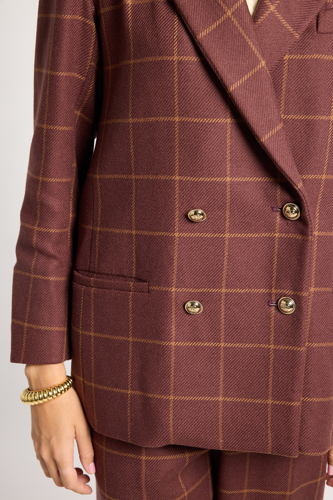 A close-up of a person wearing a double-breasted blazer and matching trousers in a rust-brown color with a thin tan windowpane plaid pattern. The blazer features gold-toned buttons and is shown against a light gray background.