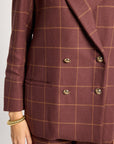 A close-up of a person wearing a double-breasted blazer and matching trousers in a rust-brown color with a thin tan windowpane plaid pattern. The blazer features gold-toned buttons and is shown against a light gray background.