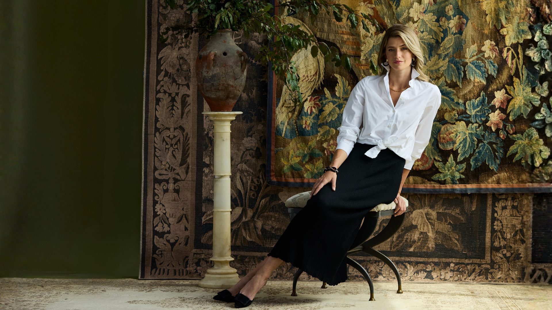 A woman sits on a chair in an ornate interior, wearing a white button-up shirt tied at the waist and a long black skirt. The background features a large tapestry and a decorative column.