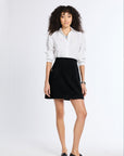A woman modeling a white long-sleeve button-up shirt and a black mini skirt, paired with black loafers, against a plain white studio background.