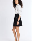 A woman with curly dark hair stands in a studio wearing a white button-up shirt, a black A-line cashmere skirt, and black flats against a plain light background.