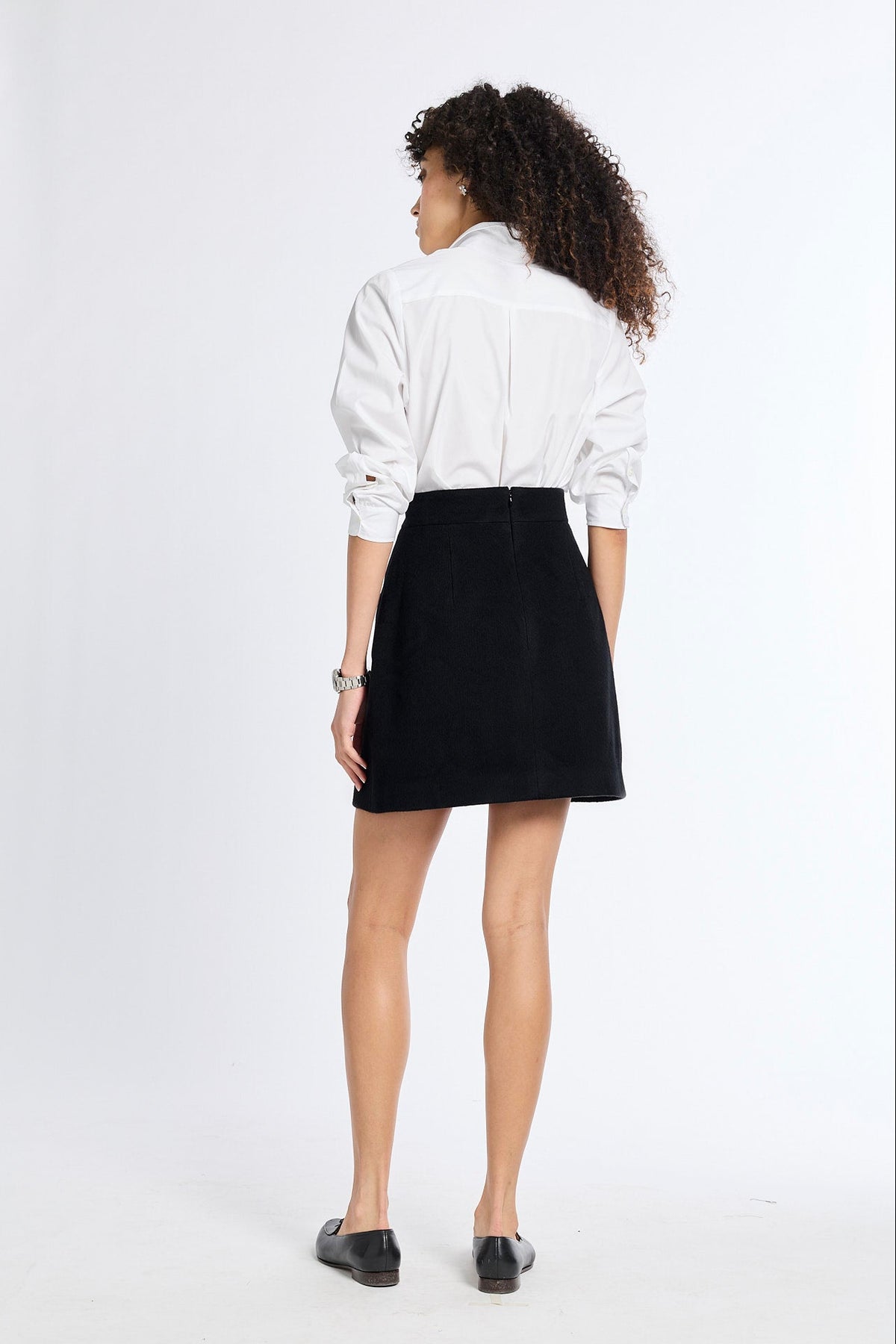 A woman is shown from behind wearing a white long-sleeve button-up shirt and a black mini skirt, paired with black flats, against a plain white studio background.
